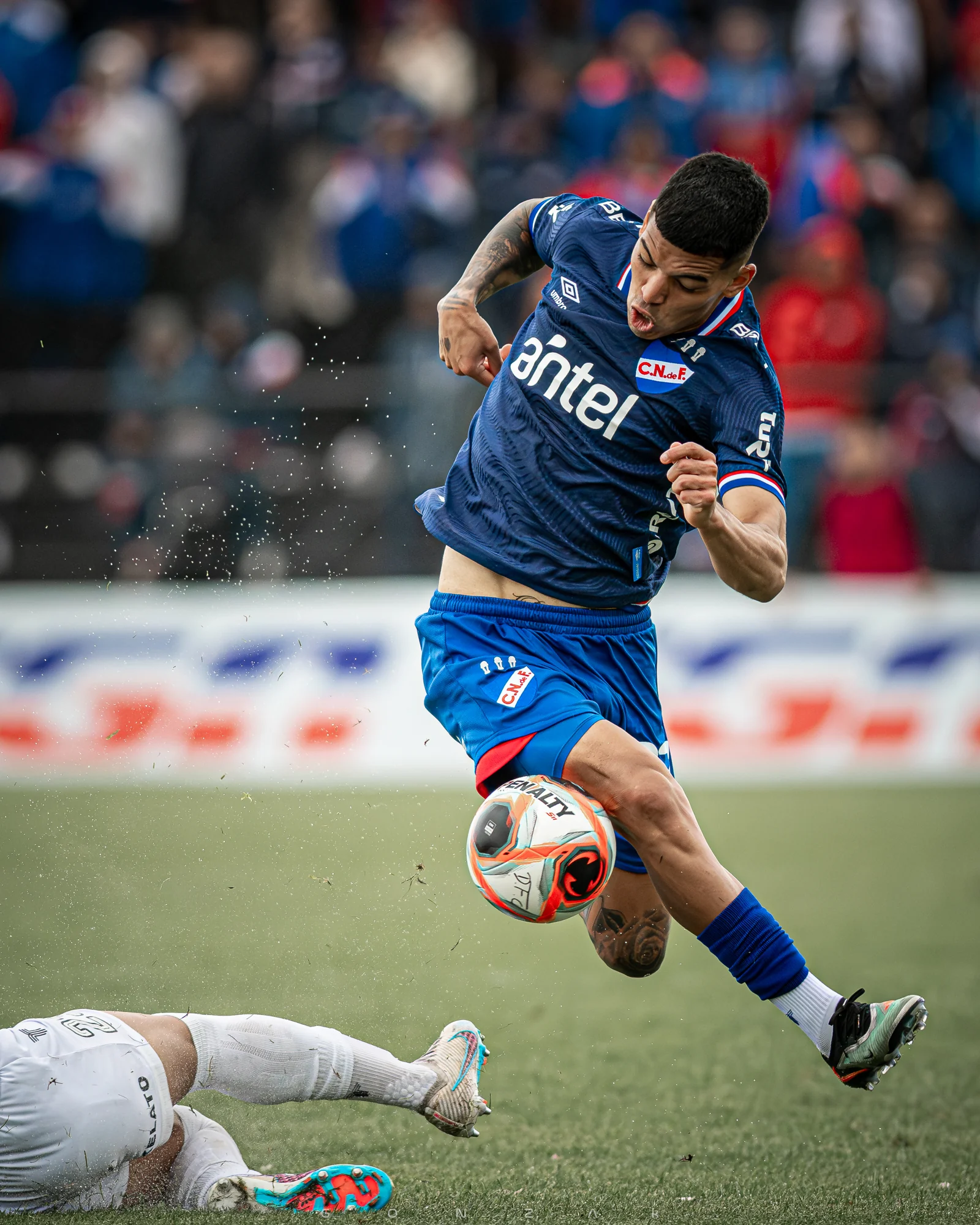 Un jugador de Nacional salta junto a la pelota mientras un rival cae al césped.