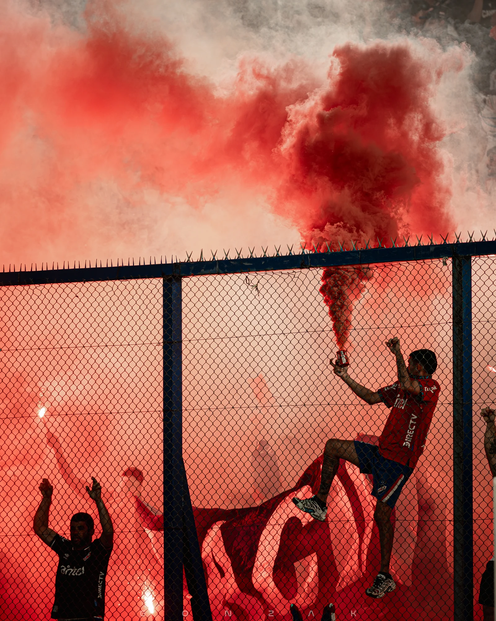 Siluetas de hinchas detrás de un alambrado entre humo rojo.