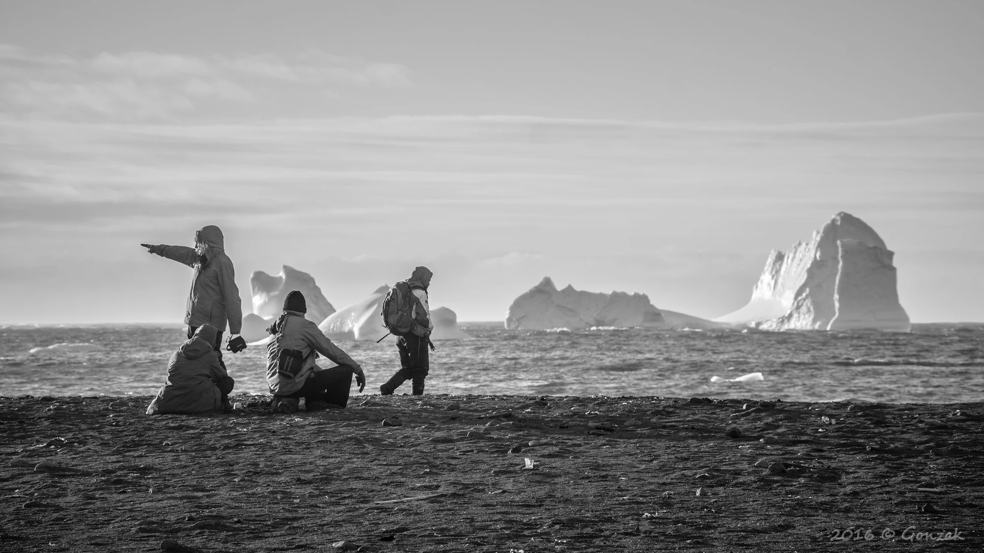 Personas recorren una costa frente a un gran iceberg en blanco y negro.